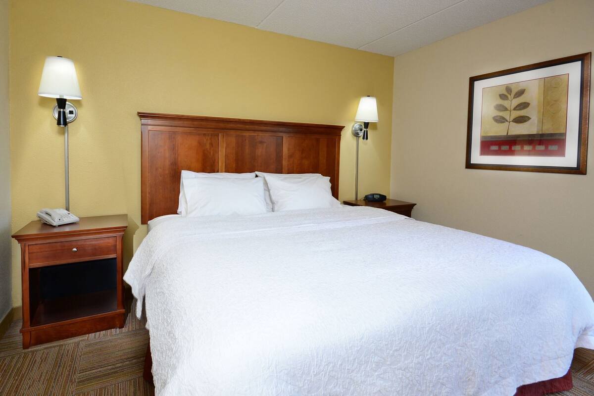 A hotel room with a large bed, two bedside tables, a nightstand, a lamp, a framed picture on the wall, and a telephone on the nightstand.