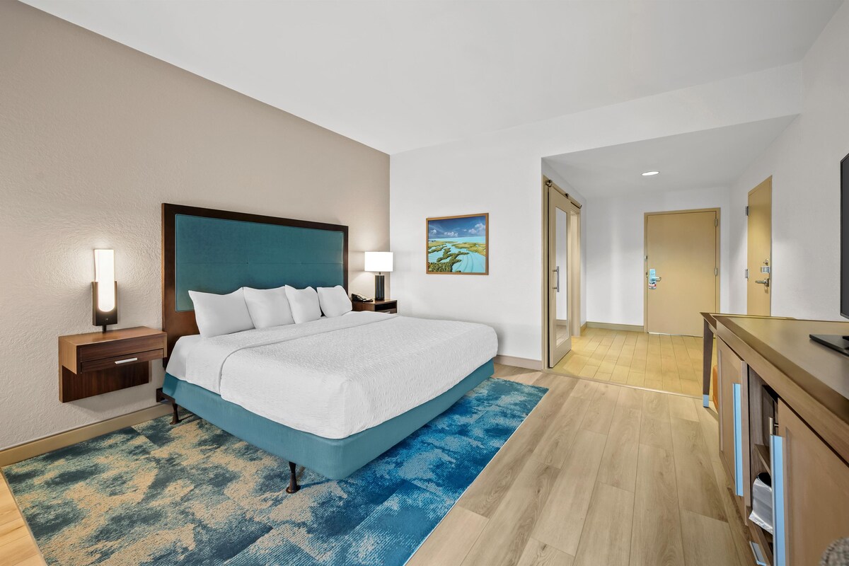A hotel room with a bed, bedside tables, a lamp, a painting, a mirror, and a TV.