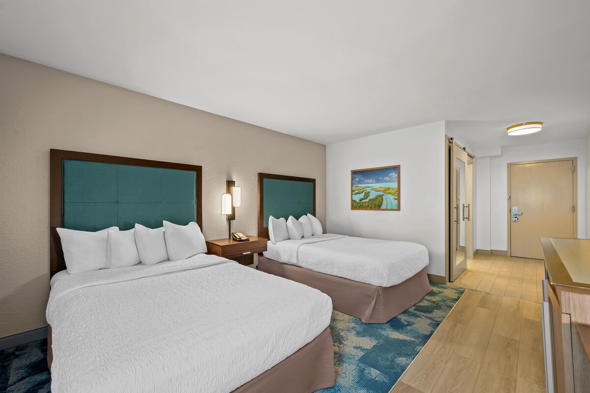 A hotel room with two beds, a nightstand with a lamp, a painting on the wall, and a door leading to another room.
