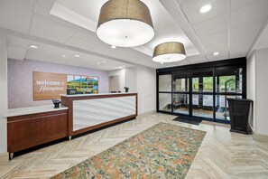 A hotel reception area with a reception desk, a welcome sign, a large window with a view, and a patterned rug.