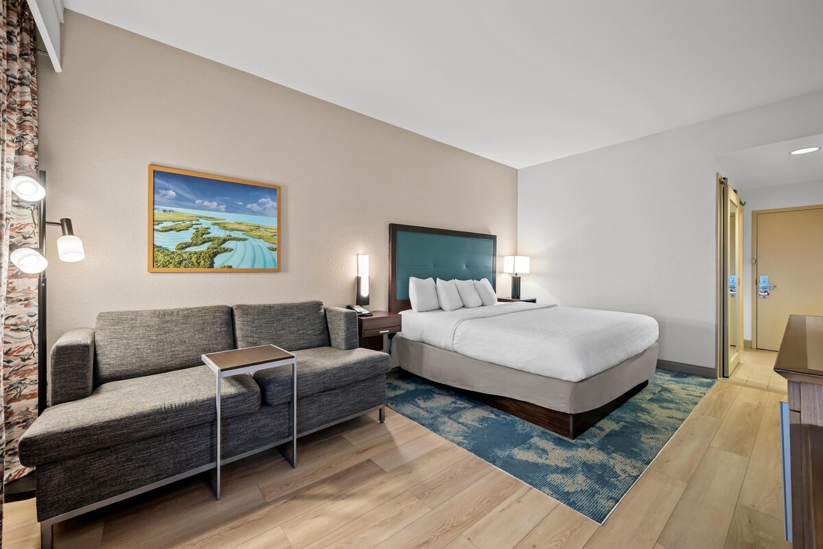 A hotel room with a bed, a sofa, a small table, a painting on the wall, and a door.