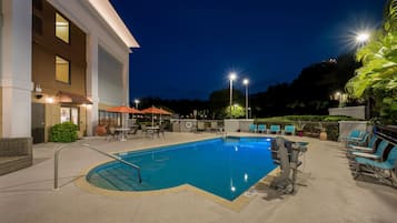 A swimming pool with lounge chairs and umbrellas, surrounded by a building with large windows and a well-lit patio area.