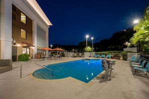 A swimming pool with lounge chairs and umbrellas, surrounded by a building with large windows and a well-lit patio area.