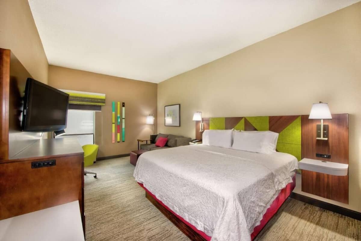 A hotel room with a bed, a desk with a television, a chair, a nightstand, and a wall-mounted lamp.