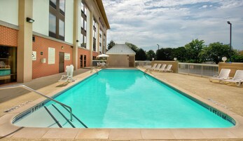 A rectangular swimming pool with a clear blue water, surrounded by a brick building with a white roof. There are lounge chairs and a small covered area with a sign indicating no diving allowed.