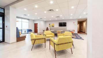 A spacious lobby with a seating area featuring yellow patterned chairs, a flat-screen TV mounted on the wall, and a large window providing natural light.