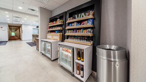 A hotel lobby with a reception desk, refrigerated display cases, shelves with snacks, and a trash bin. - Hampton Inn Winchester N/Conference Center, VA (Winchester)