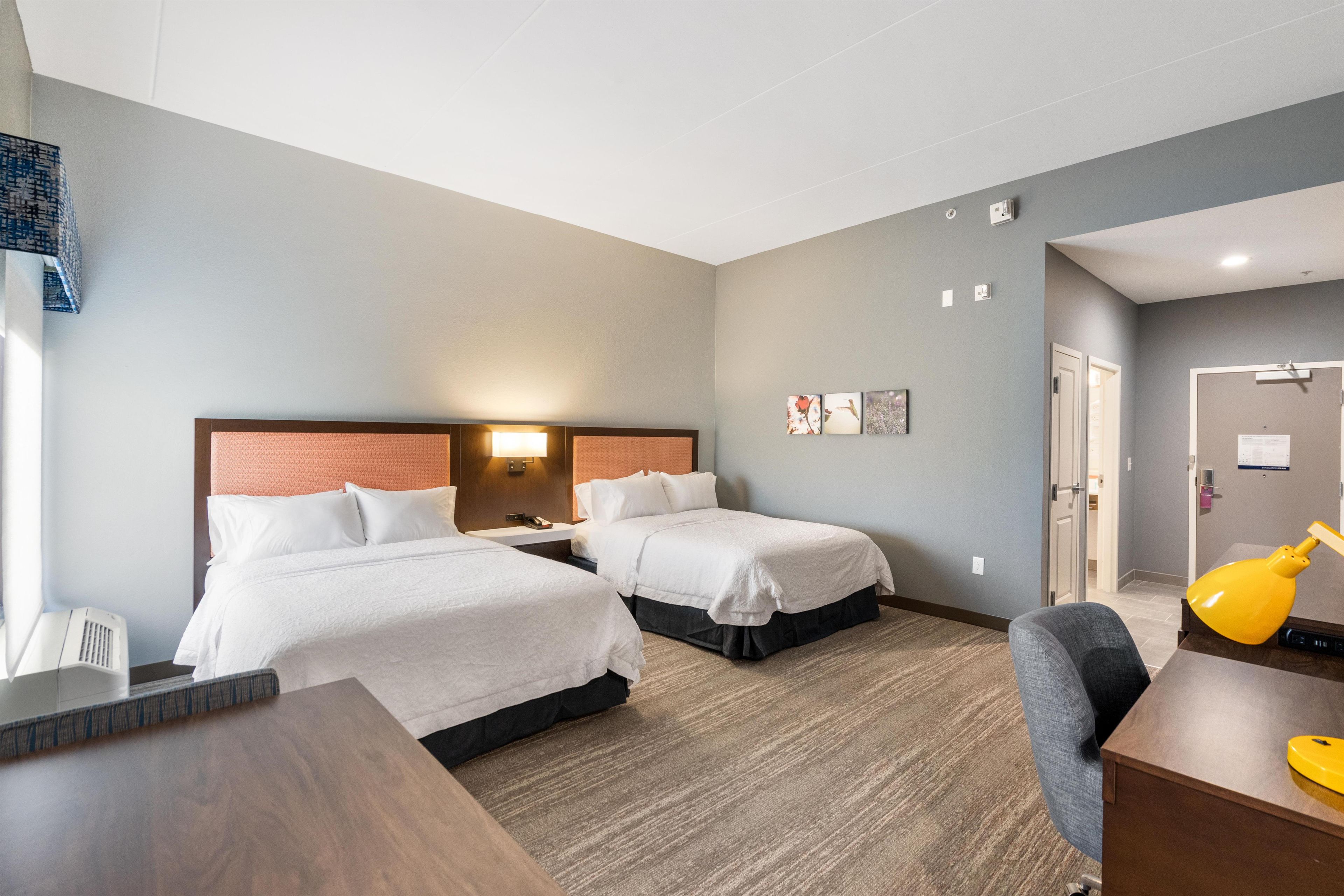 A hotel room with two beds, a desk, a chair, and a lamp.