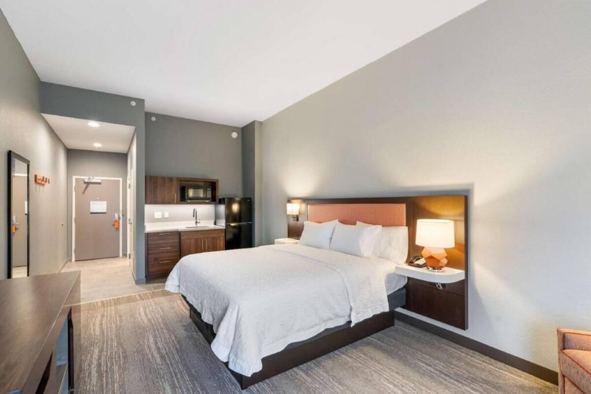 A hotel room with a bed, bedside table, lamp, and a kitchenette in the background.