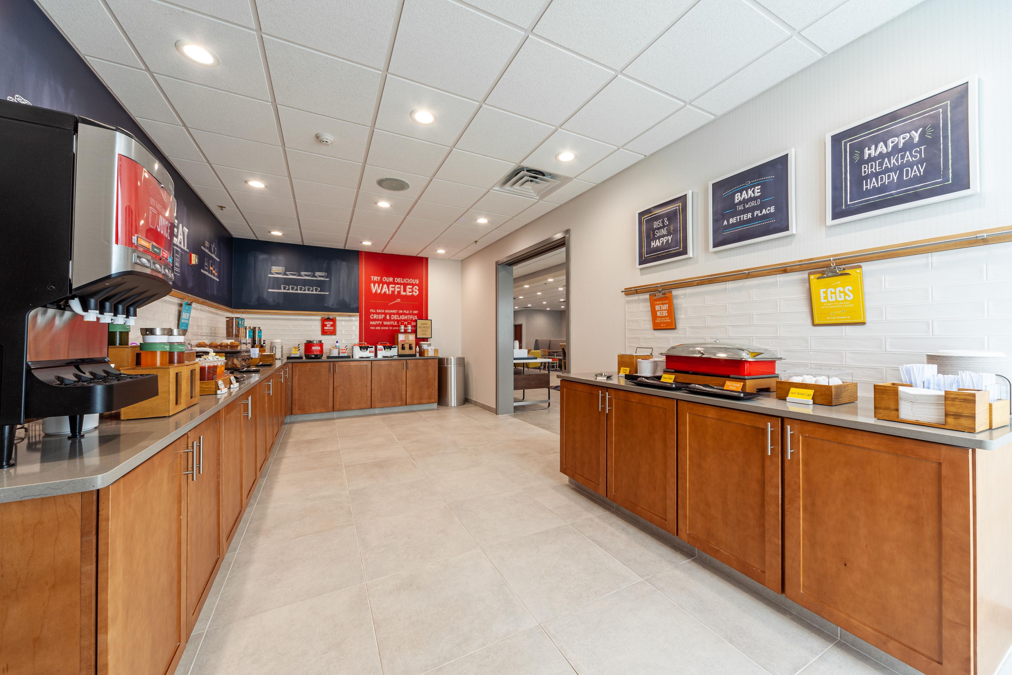 Un área de cocina moderna con una cafetera, estación de gofres y diversos alimentos exhibidos.