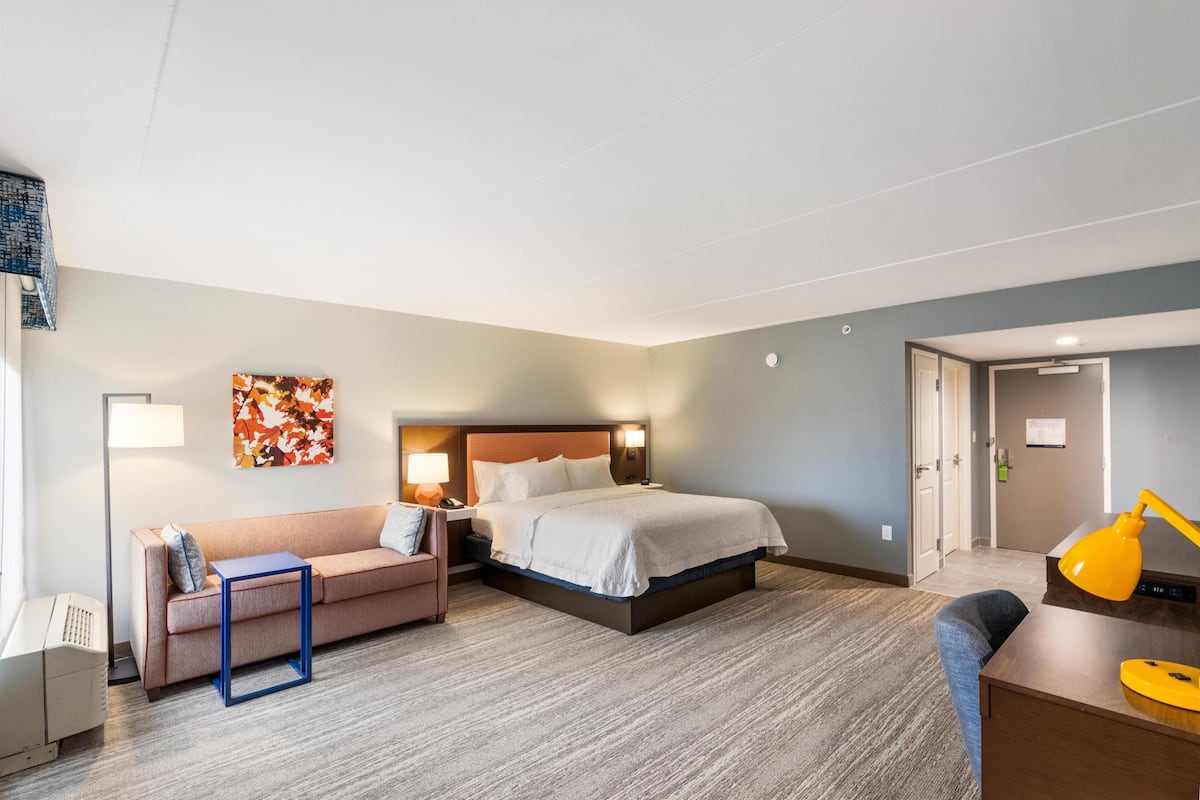 A hotel room with a bed, a sofa, a desk, a lamp, and a painting on the wall.