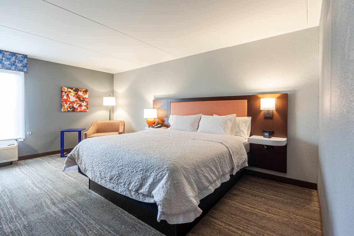 A hotel room with a large bed, two bedside tables with lamps, a small side table, and a colorful artwork on the wall.