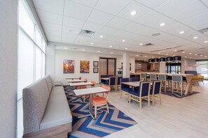 A modern dining area with tables and chairs, a seating area with a bench, and a kitchen in the background. - Hampton Inn Winchester N/Conference Center, VA (Winchester)