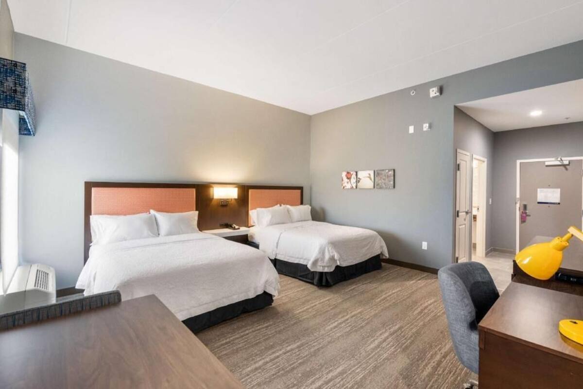 A hotel room with two beds, a desk, a chair, a lamp, and a door.