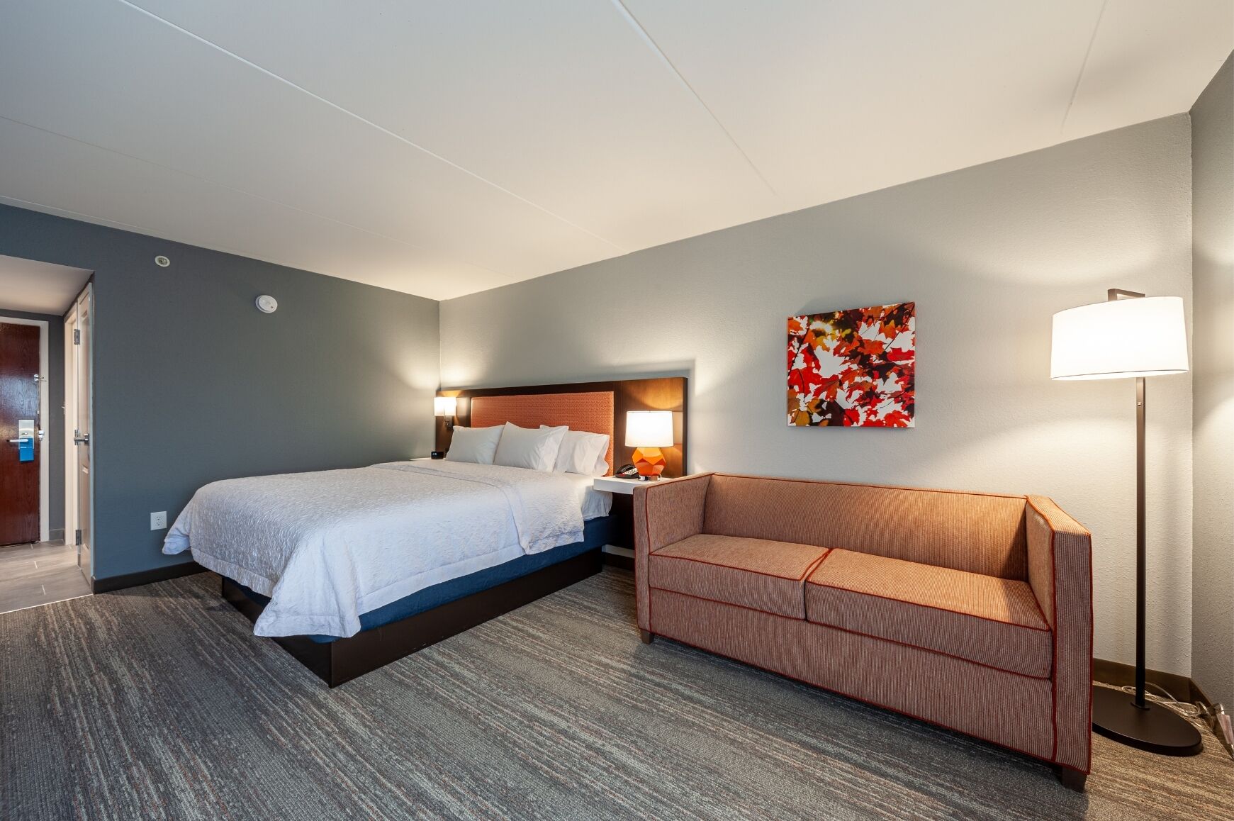 A hotel room with a bed, a sofa, a lamp, and a wall art piece.