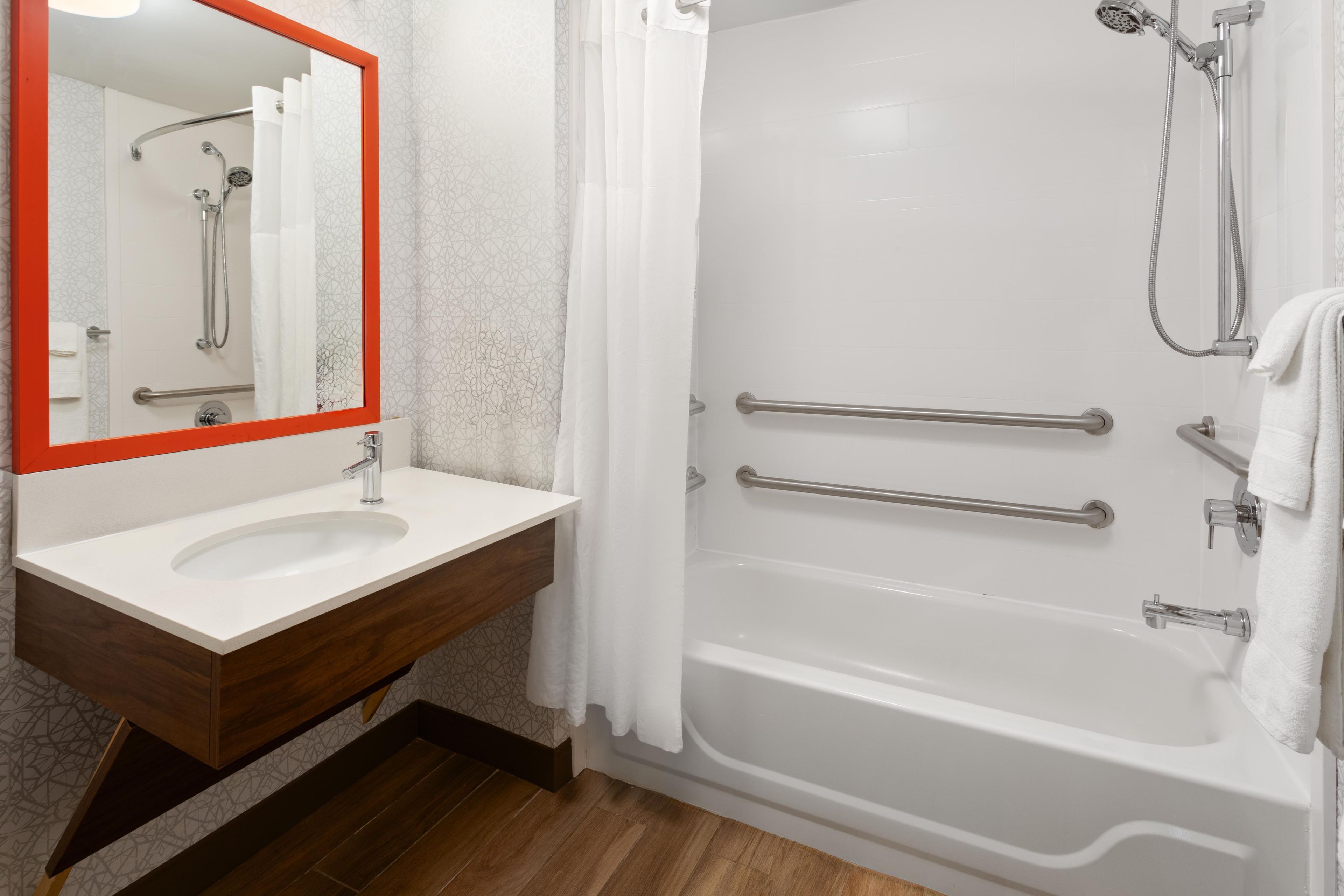 A bathroom with a white sink, a mirror with an orange frame, a shower with a handheld showerhead, and a bathtub with grab bars.