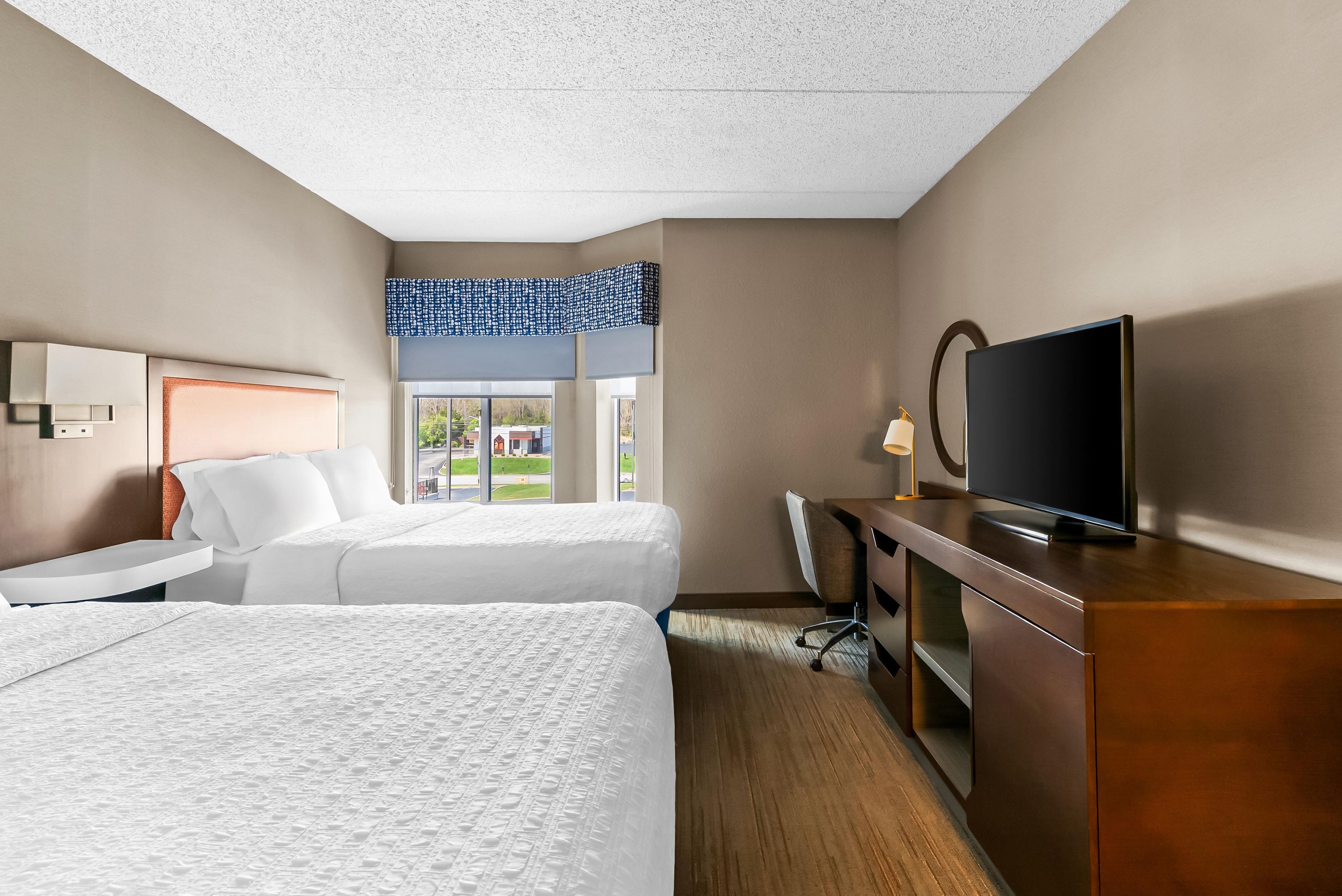 A hotel room with two beds, a desk, a television, and a window with a view of the outside.