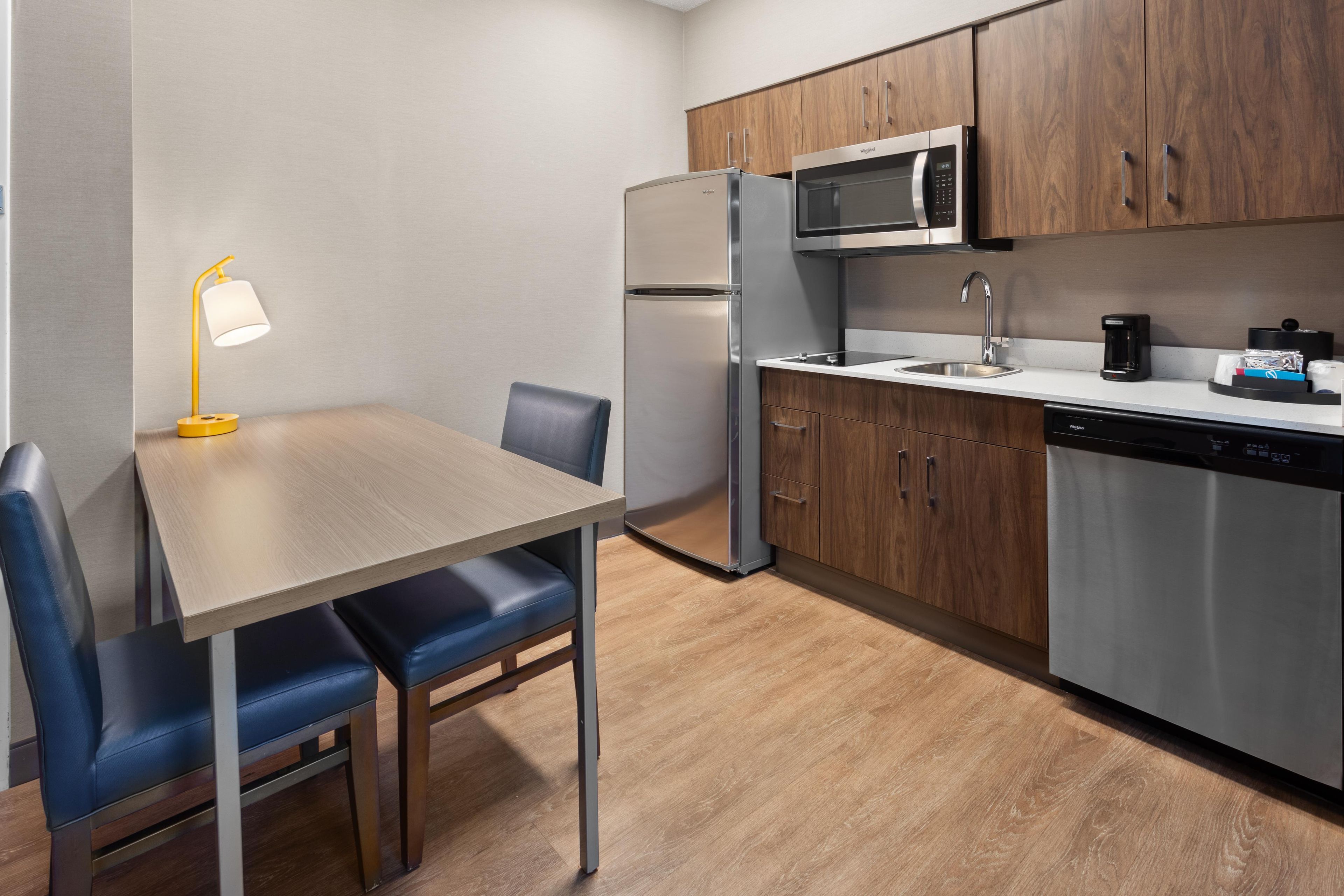 A compact kitchen with a microwave, refrigerator, and dishwasher. There is a dining table with chairs and a small lamp.