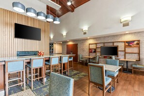 A modern bar area with a flat-screen TV, wooden panel walls, and blue upholstered chairs. - Hampton Inn & Suites Scottsburg (Scottsburg)