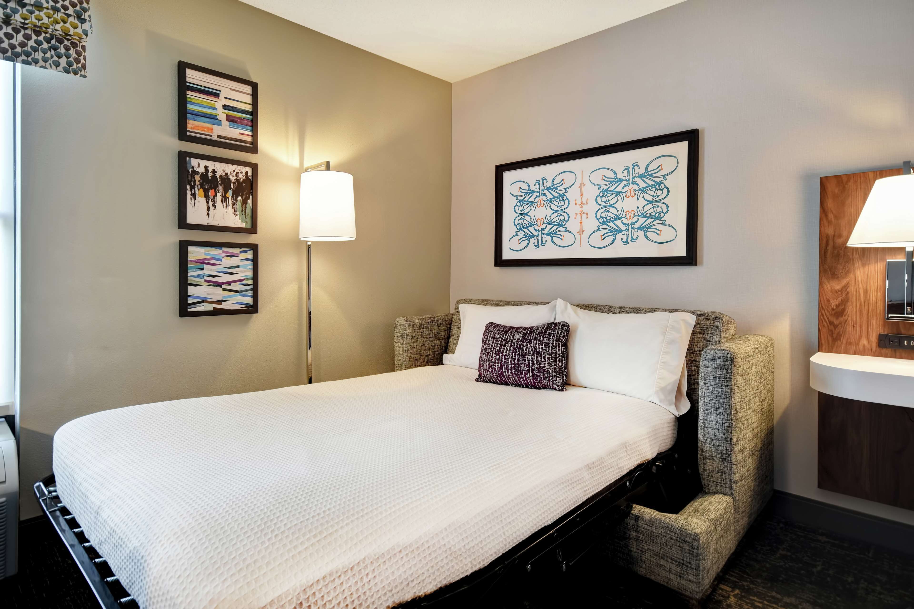 A neatly made bed with a white comforter and two pillows, a wall-mounted lamp, and framed artwork on the wall.