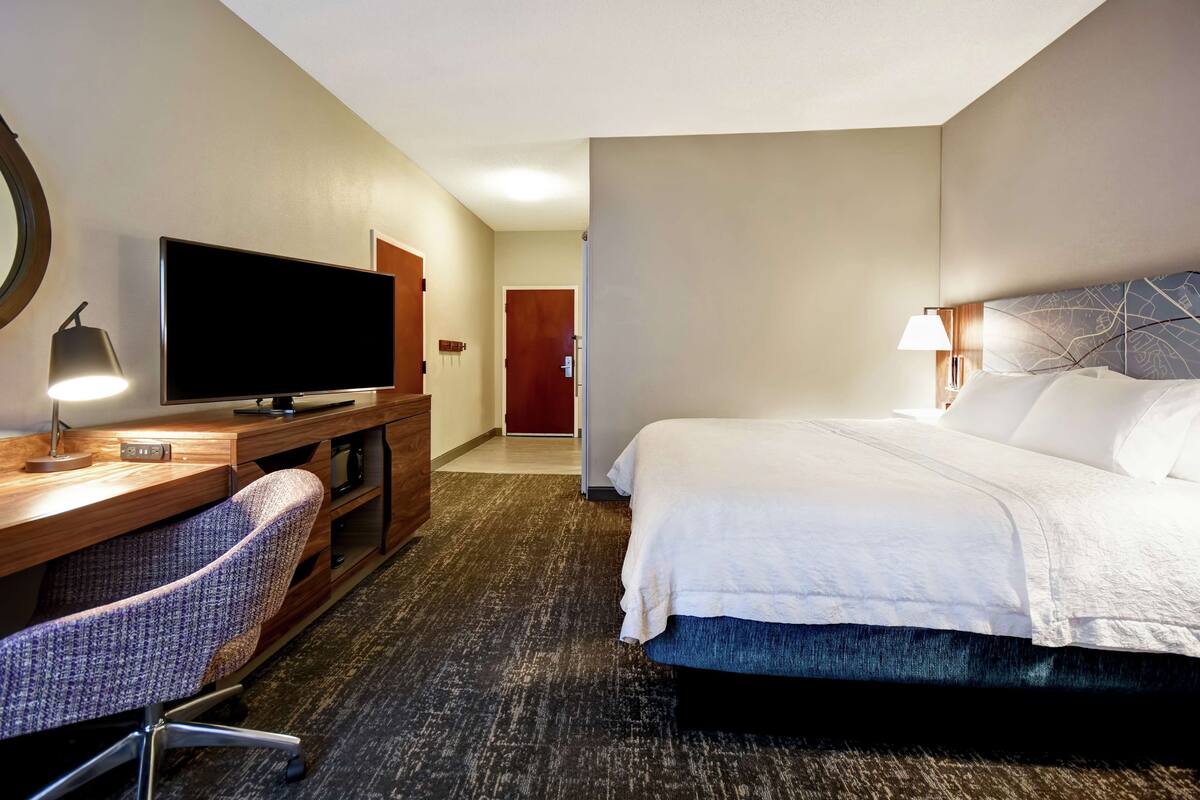 A hotel room with a bed, a desk with a television, a chair, and a lamp.