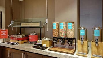 Un comptoir pour le petit-déjeuner avec différents distributeurs de nourriture, un panneau d’avertissement indiquant que certaines surfaces sont chaudes, et un panneau indiquant que le petit-déjeuner commence ici.