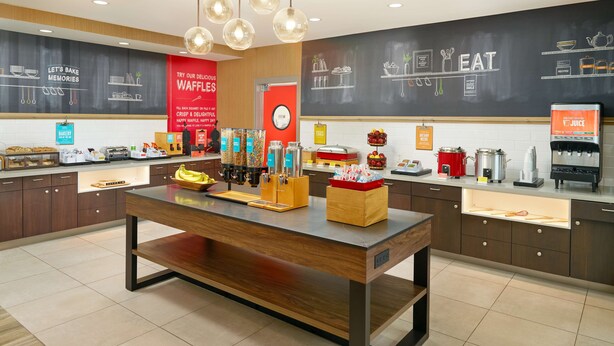 Um café moderno com uma mesa de madeira central, diversos itens alimentares e um menu em lousa.