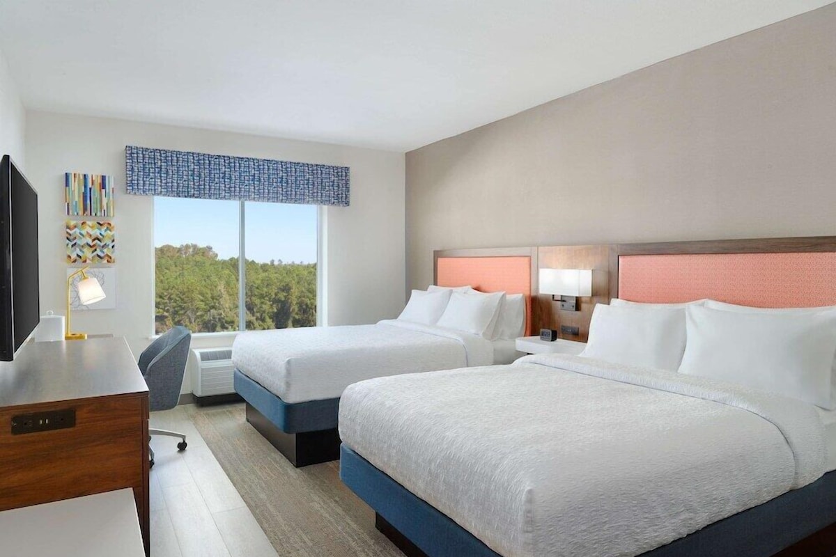 A hotel room with two beds, a desk, a TV, and a large window with a view of trees.