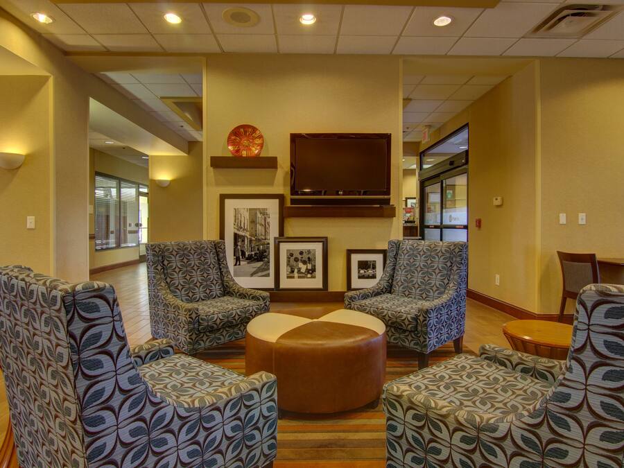 A lobby with patterned armchairs, a central ottoman, framed pictures on the wall, and a flat-screen TV.