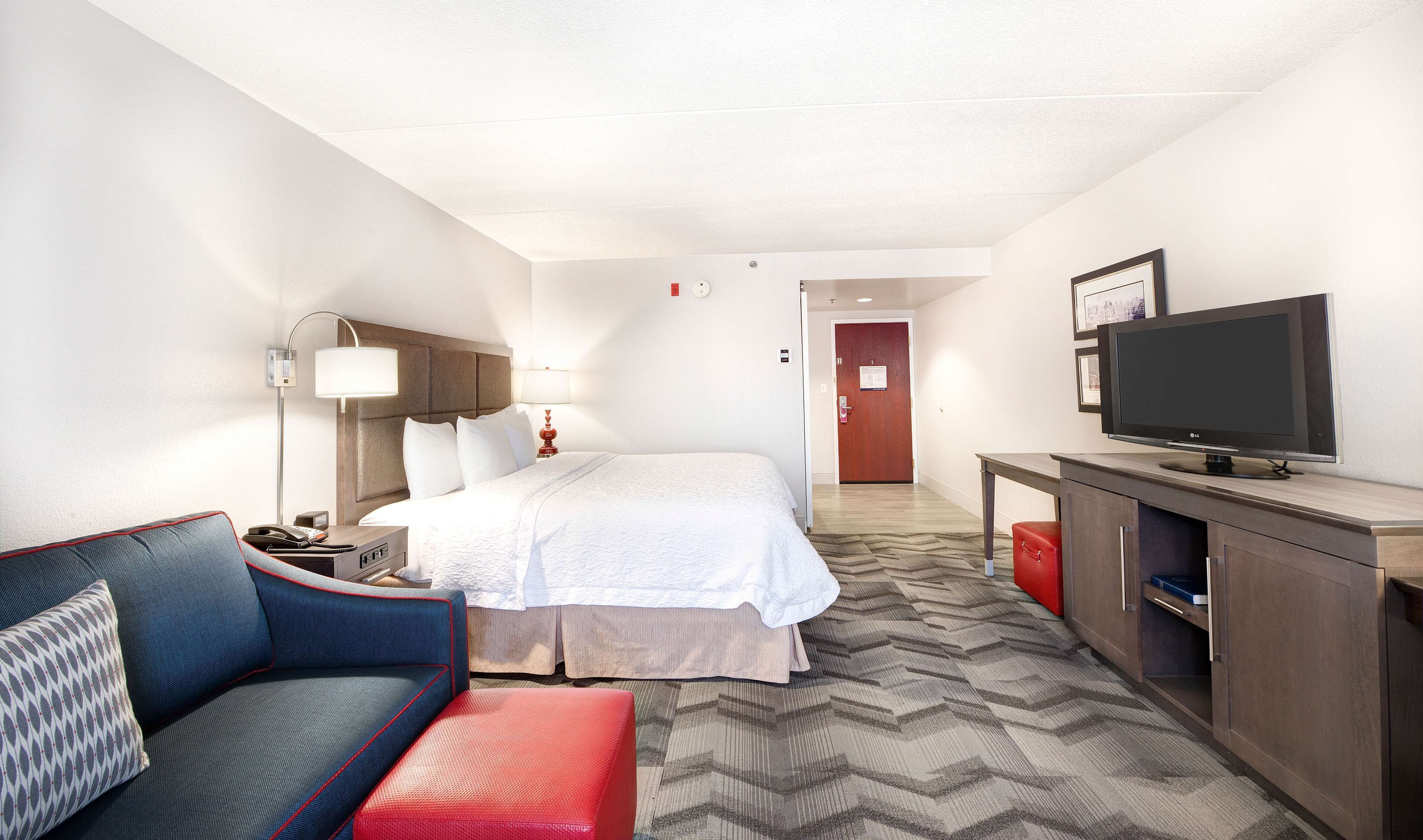 A hotel room with a bed, a TV, a desk, a lamp, and a sofa.