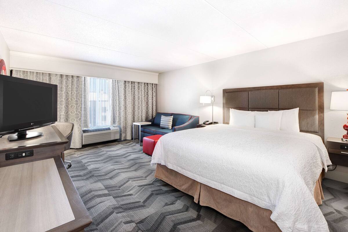 A hotel room with a large bed, a television, a desk with a computer, a sofa, and a window with curtains.