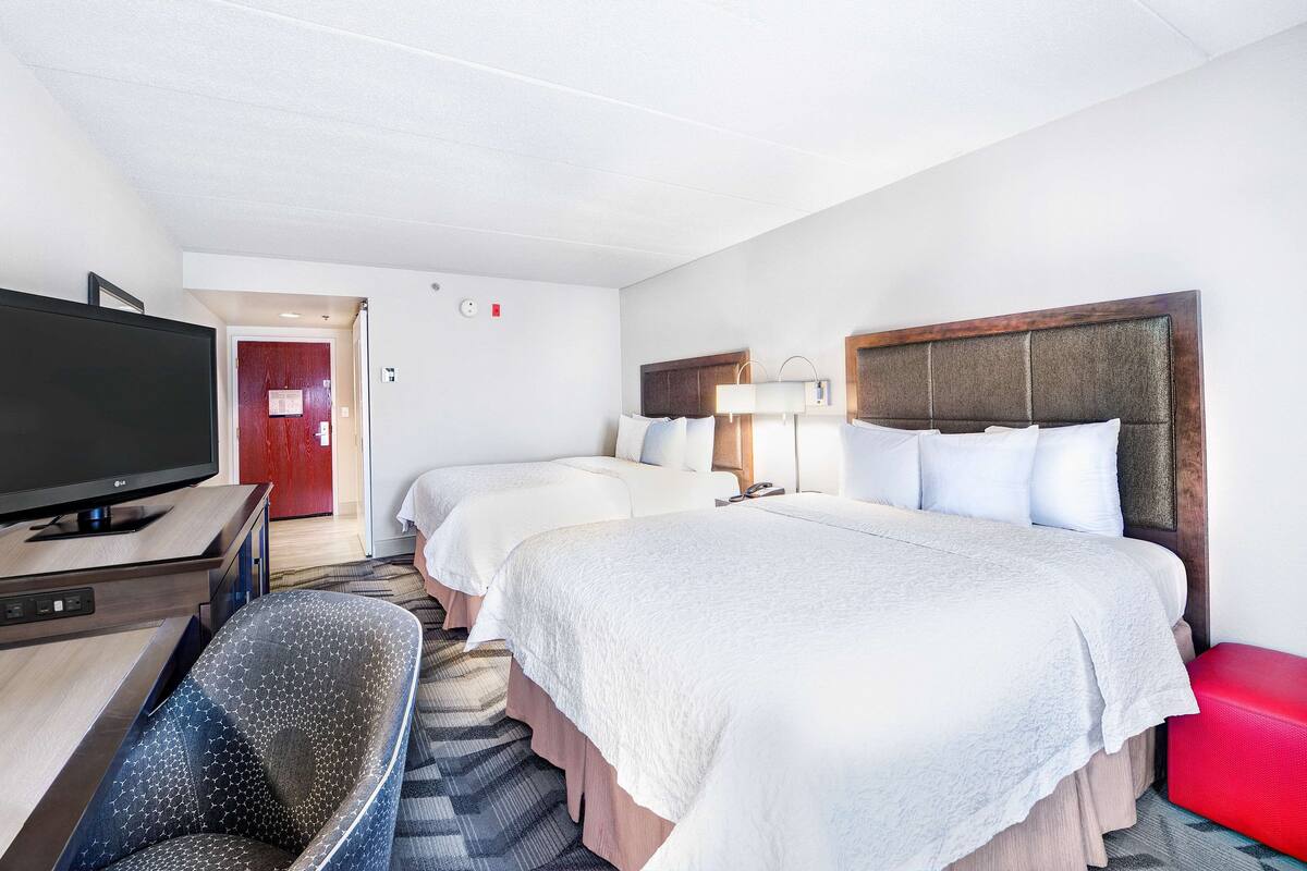 A hotel room with two beds, a TV, a desk, and a red ottoman.