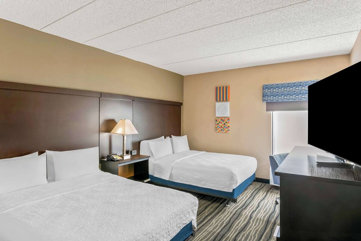A hotel room with two beds, a desk, a lamp, and a window with blinds.