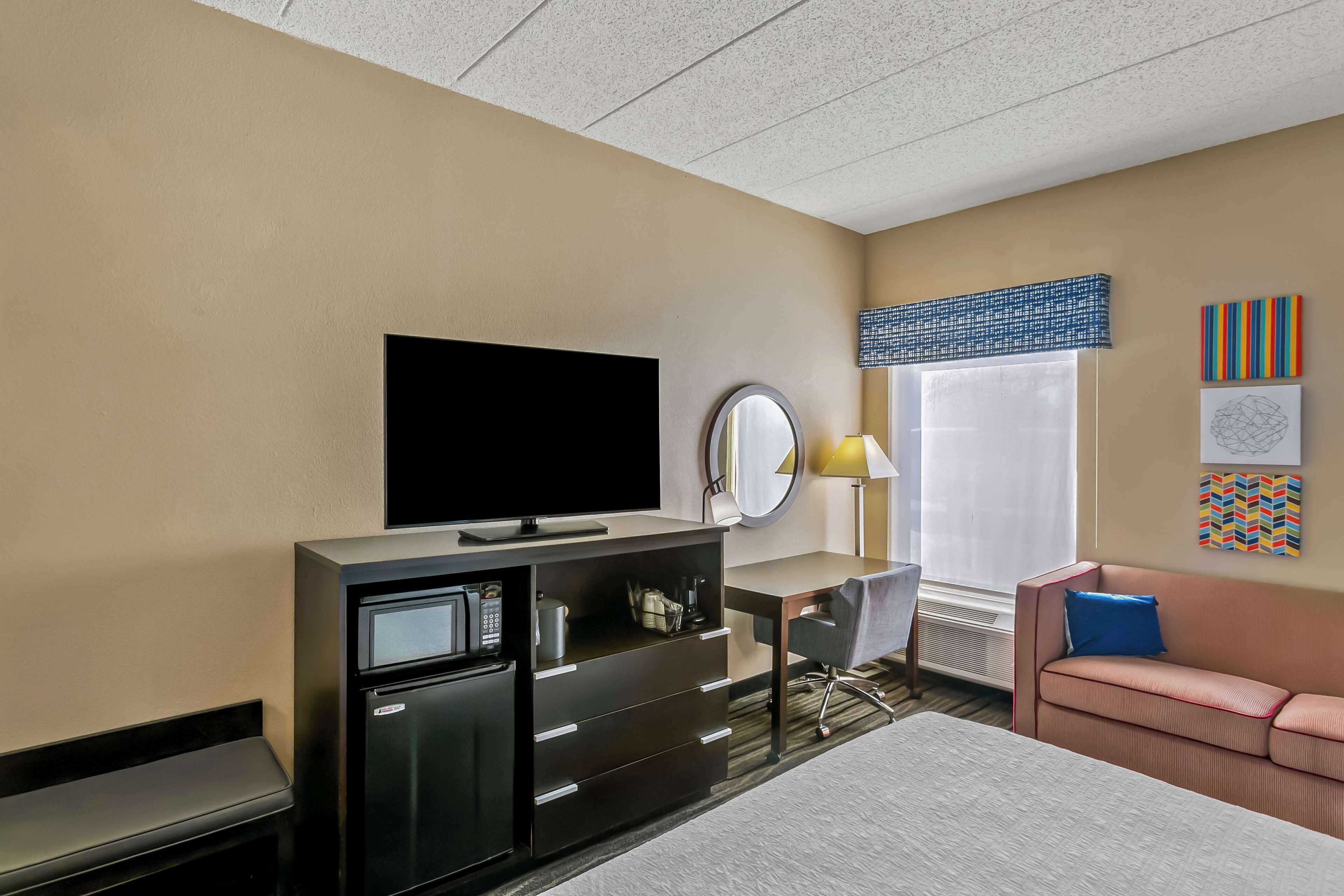 A hotel room with a flat-screen TV, a minibar, a desk, a chair, a sofa, a window with blinds, and artwork on the wall.