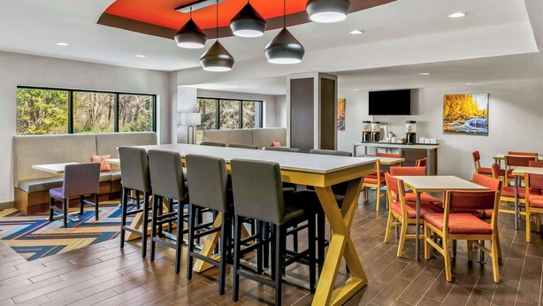 Un comedor moderno con varias mesas y sillas, una zona de cocina con televisor de pantalla plana, y amplias ventanas que ofrecen vistas al exterior.
