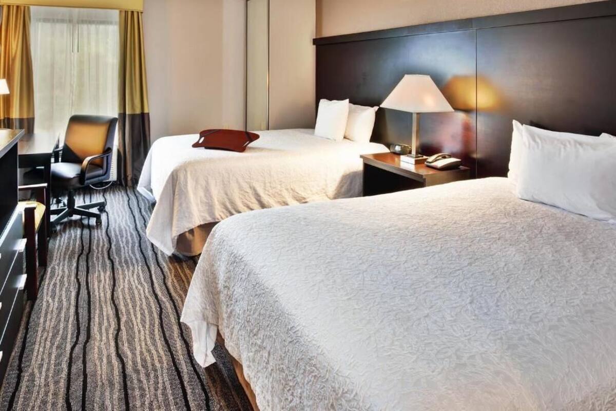 Habitación de hotel con dos camas, un escritorio, una silla, una lámpara y un ventanal con cortinas.