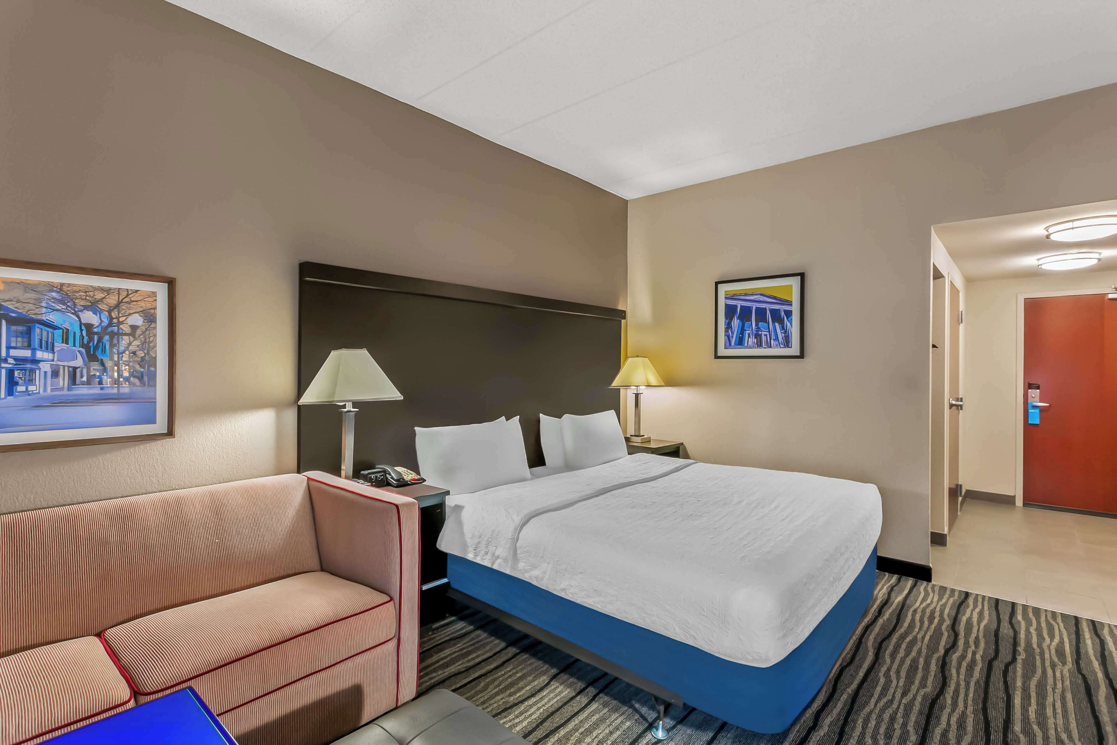A hotel room with a bed, a sofa, two bedside lamps, a framed picture on the wall, and a door leading to another room.