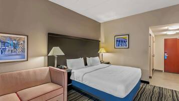 A hotel room with a bed, a sofa, two bedside lamps, a framed picture on the wall, and a door leading to another room.
