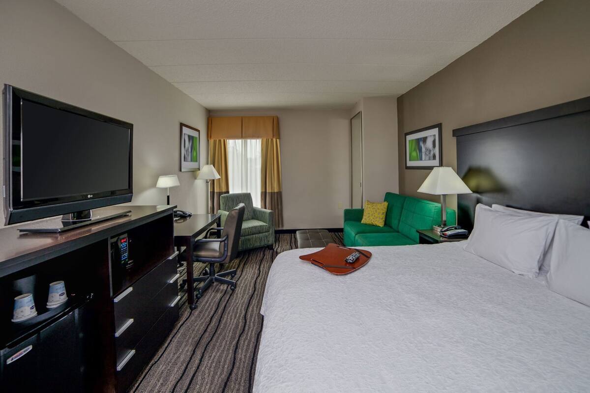 A hotel room with a bed, a television, a desk with a chair, a green sofa, and a lamp.