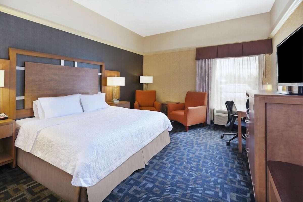 A hotel room with a large bed, two chairs, a desk, a television, and a window with curtains.