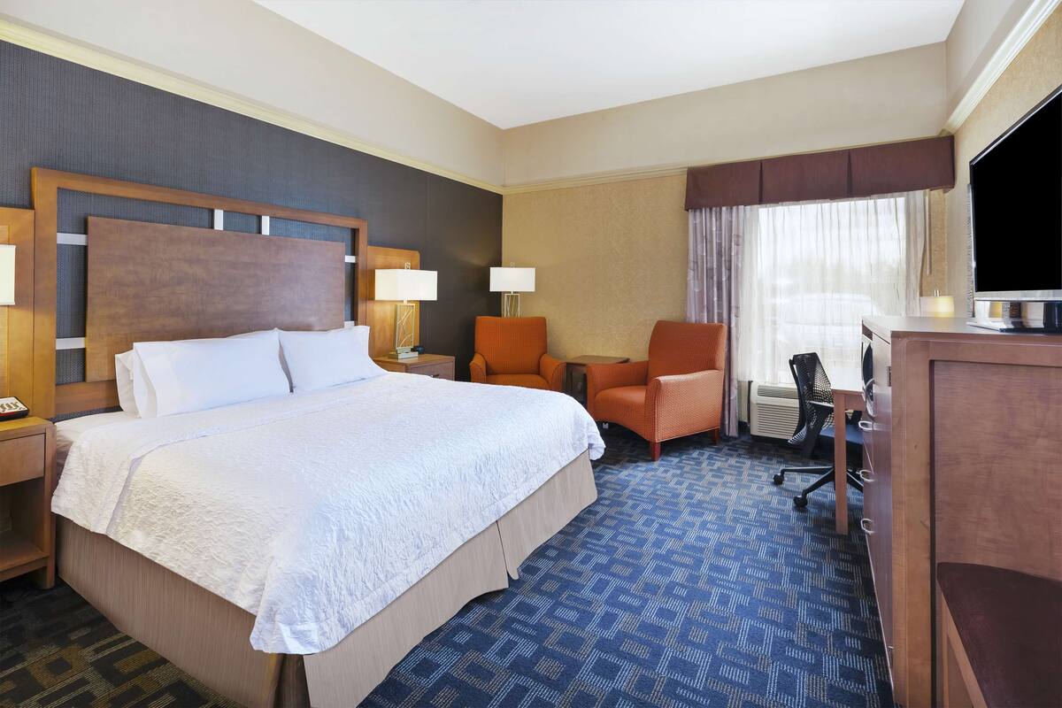 A hotel room with a large bed, two chairs, a desk, a TV, and a window with curtains.