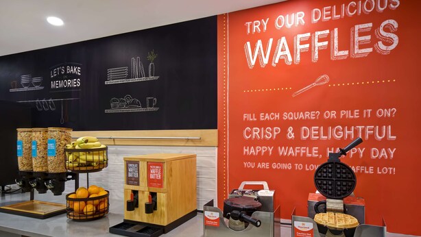 A waffle station with various waffle makers and a sign promoting waffles. - Hampton Inn Potomac Mills/Woodbridge (Woodbridge)