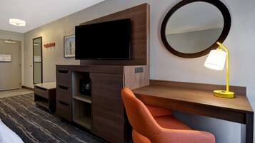 A hotel room with a wooden desk, a television, a mirror, and a lamp.