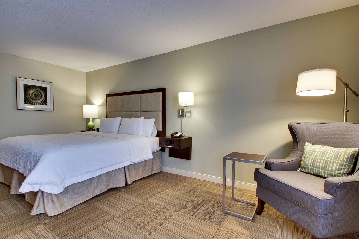 A hotel room with a bed, bedside table, lamp, chair, and a framed picture on the wall.