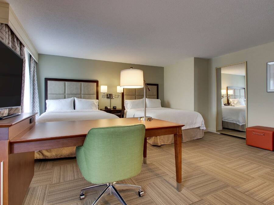 A hotel room with two beds, a desk, a green chair, a lamp, and a mirror.