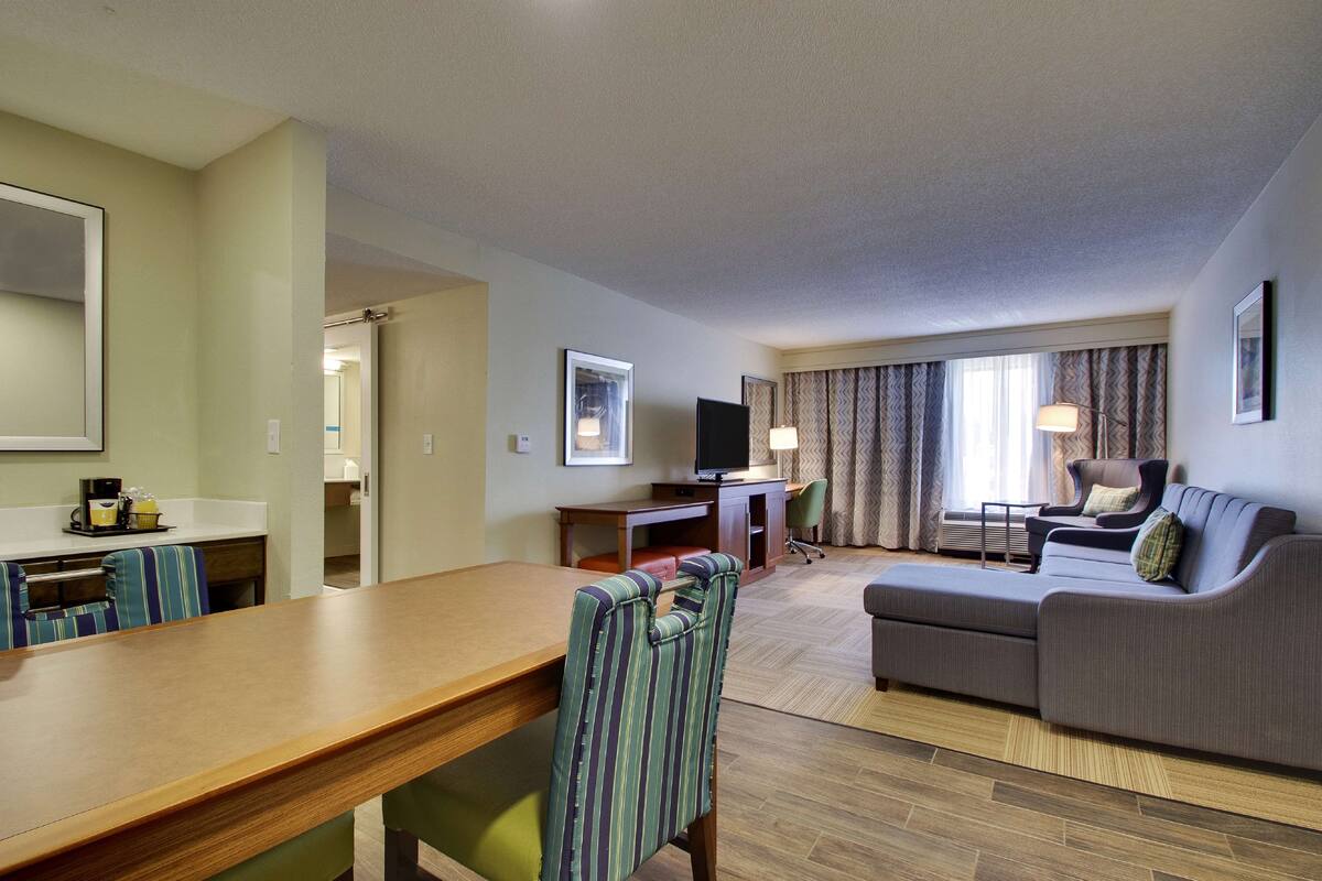 A hotel room with a wooden dining table, striped armchair, grey sofa, and a TV on a desk.