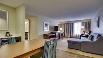 A hotel room with a wooden dining table, striped armchair, grey sofa, and a TV on a desk.