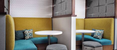 A corner seating area with a yellow and teal upholstered bench, a small round table, and a grey ottoman.