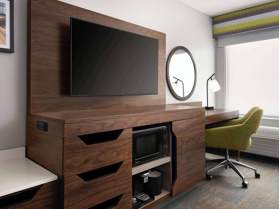Ein modernes Hotelzimmer mit einem Flachbildfernseher an einer Holzkommode, einem Schreibtisch mit Stuhl, einem runden Spiegel und einem Fenster mit Jalousien.