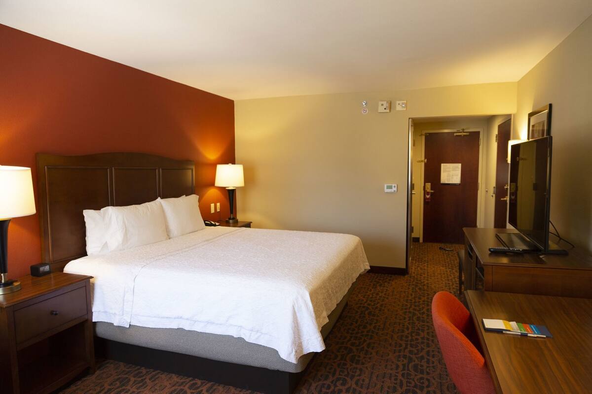 A hotel room with a large bed, bedside lamps, a desk with a computer, a chair, and a door leading to another room.